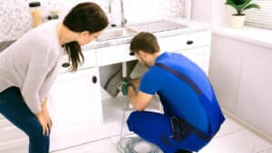 A plumber kneeling beside a woman as they work together to fix a pipe. The plumber is wearing overalls and holding a wrench plumber Plano